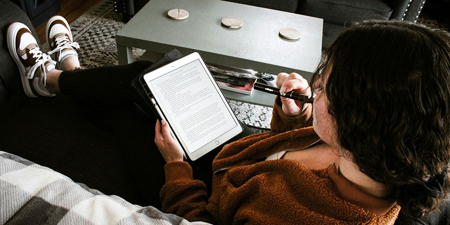 a person sitting on a couch, reading an e-book on a tablet while holding a vape device